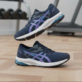 Asics blue trainers with white soles and purple and blue logo, displayed on artificial grass.