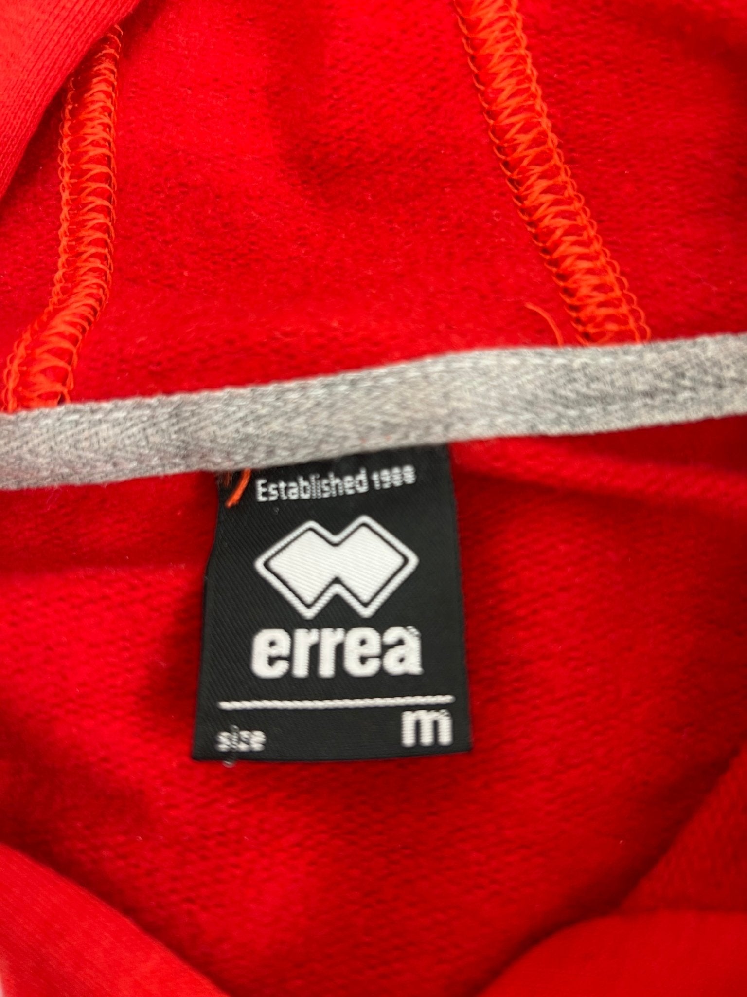 Close-up of the Errea brand label inside the Volleyball England red hoodie showing medium size
