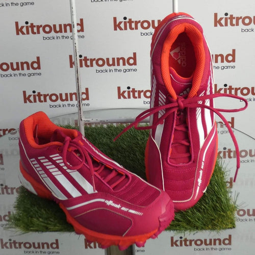 Pair of adidas pink and red hockey shoes with white stripes displayed on an artificial grass platform