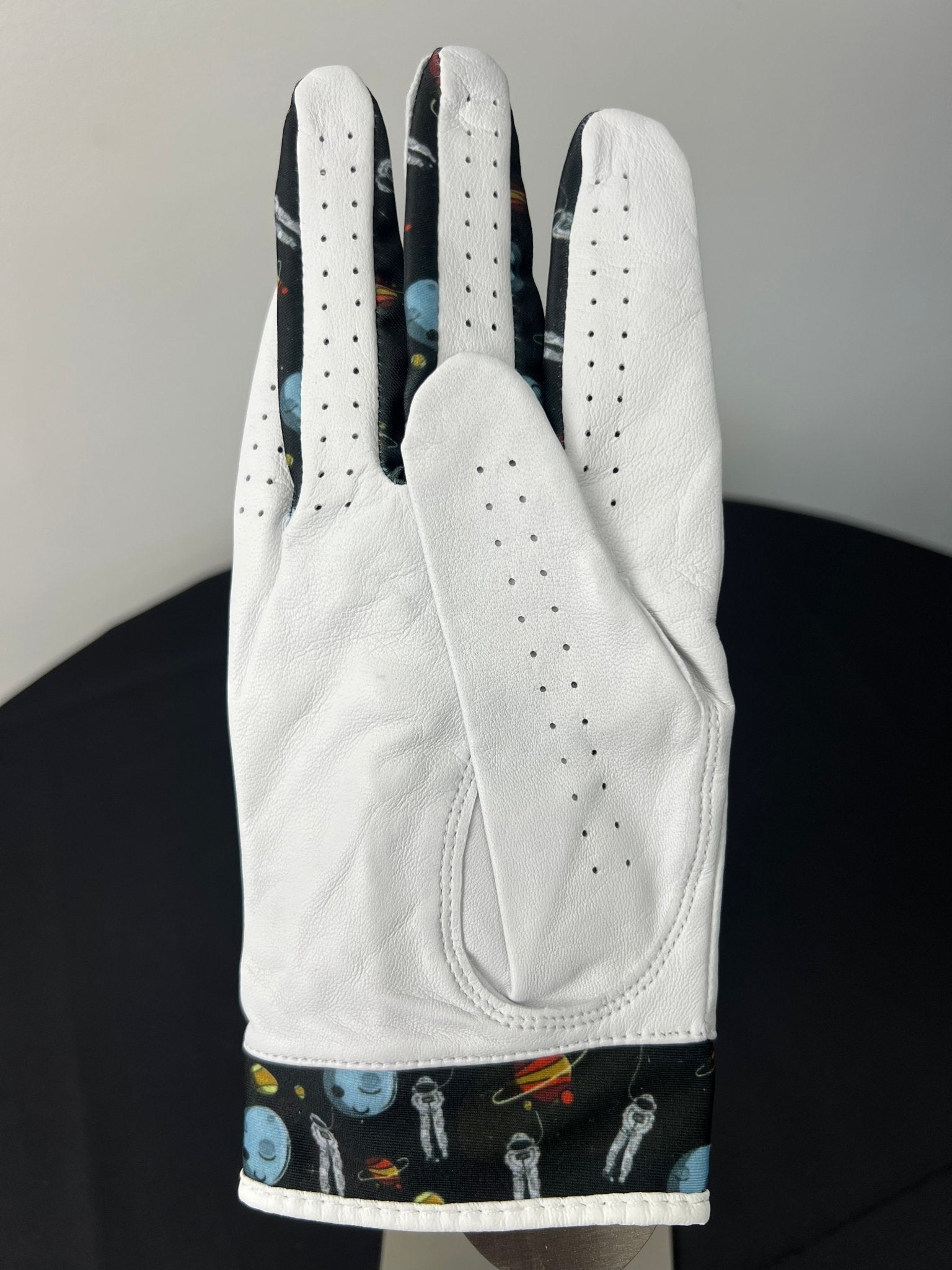 Back view of white women's golf glove showing black space-themed detailing with planets and astronauts